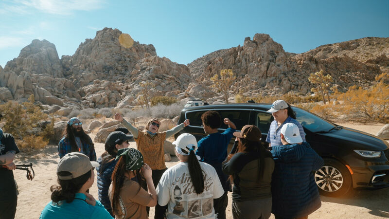 A group of people are gathered near a black minivan in a desert landscape. The backdrop features rocky mountains and sparse vegetation. Some individuals are standing with their arms raised, while others are looking towards the camera. The overall scene suggests a group outing or excursion in a natural setting.
